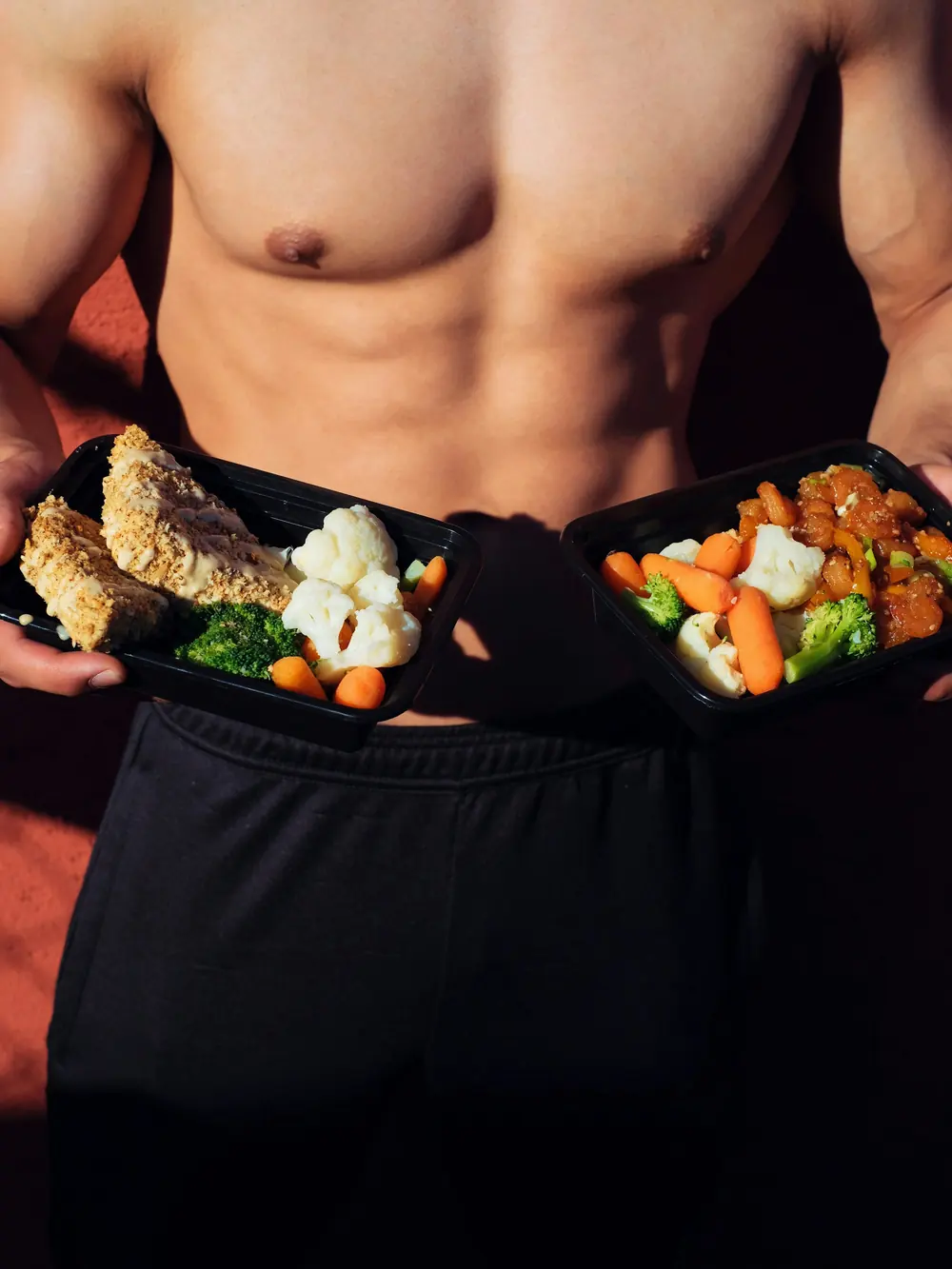 An athlete holding two meal prep containers of chicken and vegetables